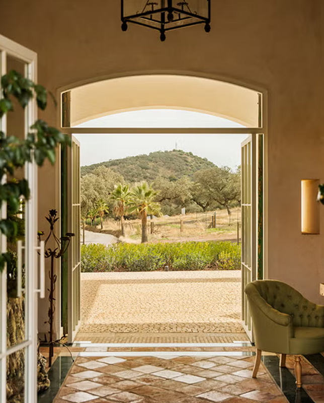 Vista desde el interior de una finca hacia colinas y palmeras, a través de grandes puertas abiertas.